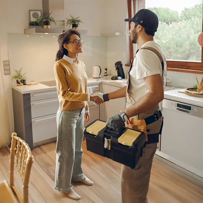 Professional repairman in uniform shaking hands with woman while standing at home kitchen.