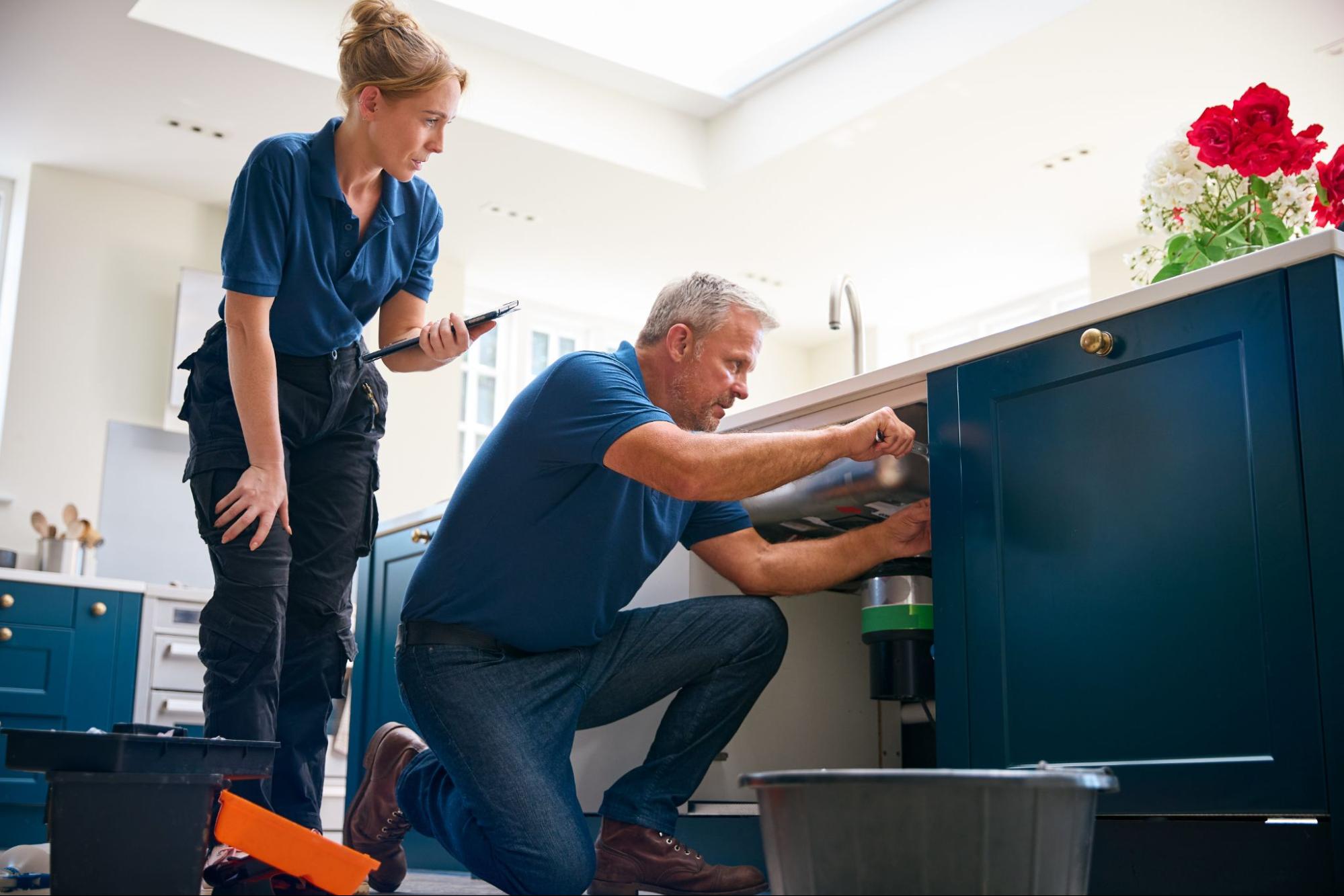 Professional home repair service team fixing kitchen sink disposal with digital tablet guidance.