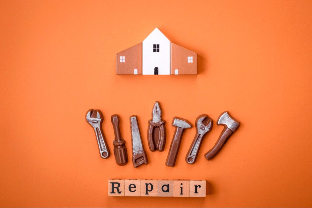 Symbolic home repair service setup with a model house and toy tools against a bright orange background.