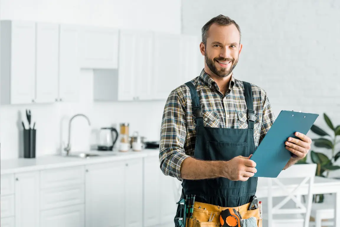 Smiling handsome plumber holding clipboard and looking at camera in kitchen.