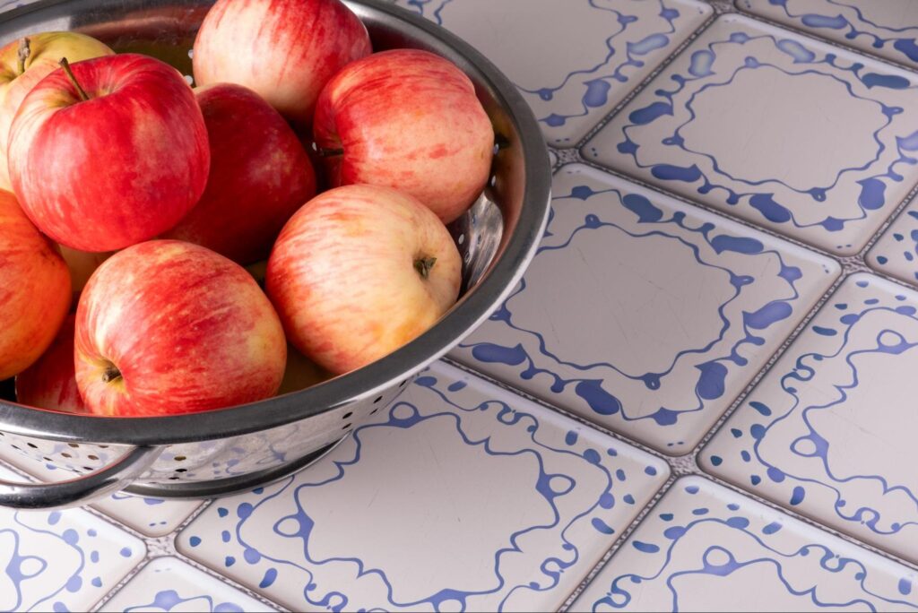 A bowl full of apples sits on a counter with white and blue kitchen tile.